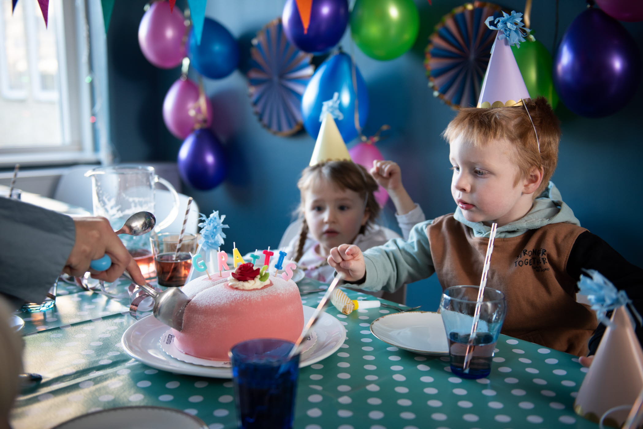 Barn i partyhattar skär upp en tårta som det står "grattis" på. I bakgrunden hänger balonger i olika färger.